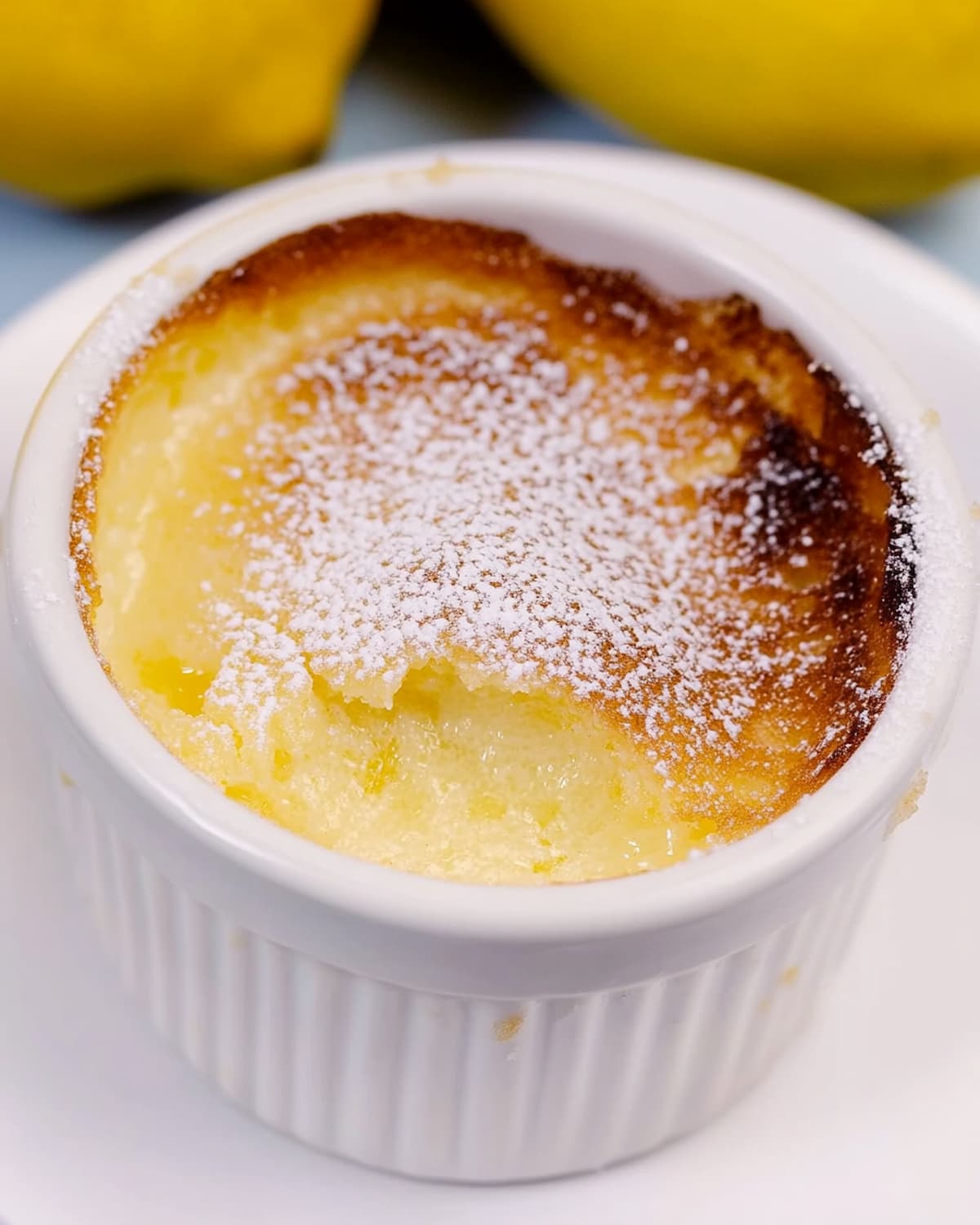 Ramequin avec un soufflé au citron au Air Fryer, doré sur le dessus, saupoudré de sucre glace, posé sur une assiette blanche avec des citrons en arrière-plan.