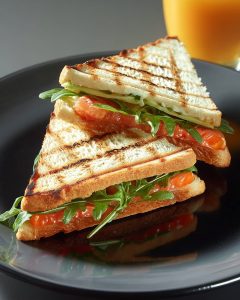 Croque monsieur au saumon au Air Fryer, grillé à la perfection, garni de roquette et tomates, servi en triangles sur une assiette noire.