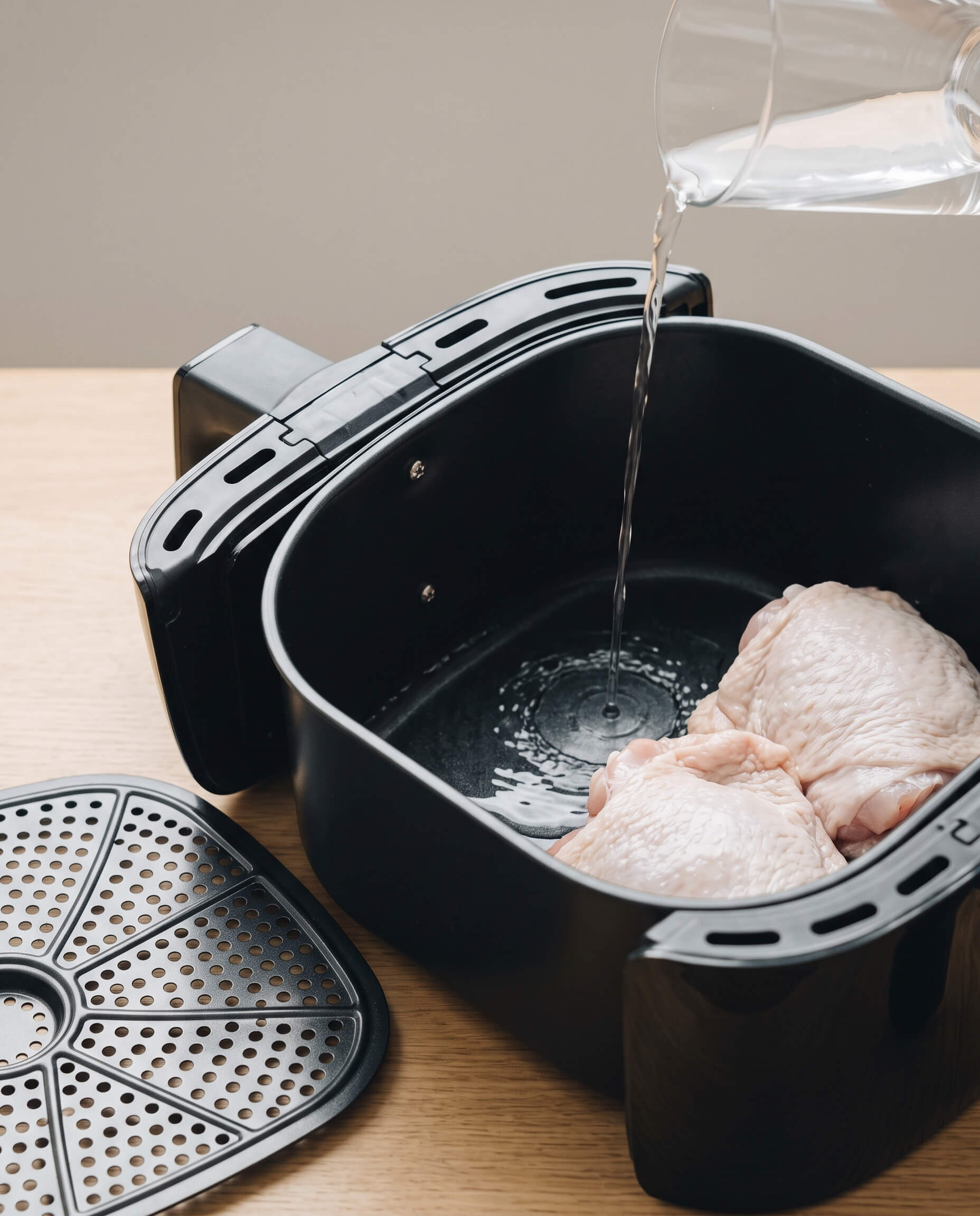 Eau versée dans le tiroir d'un air fryer, grille retirée, cuisses de poulet crues à côté