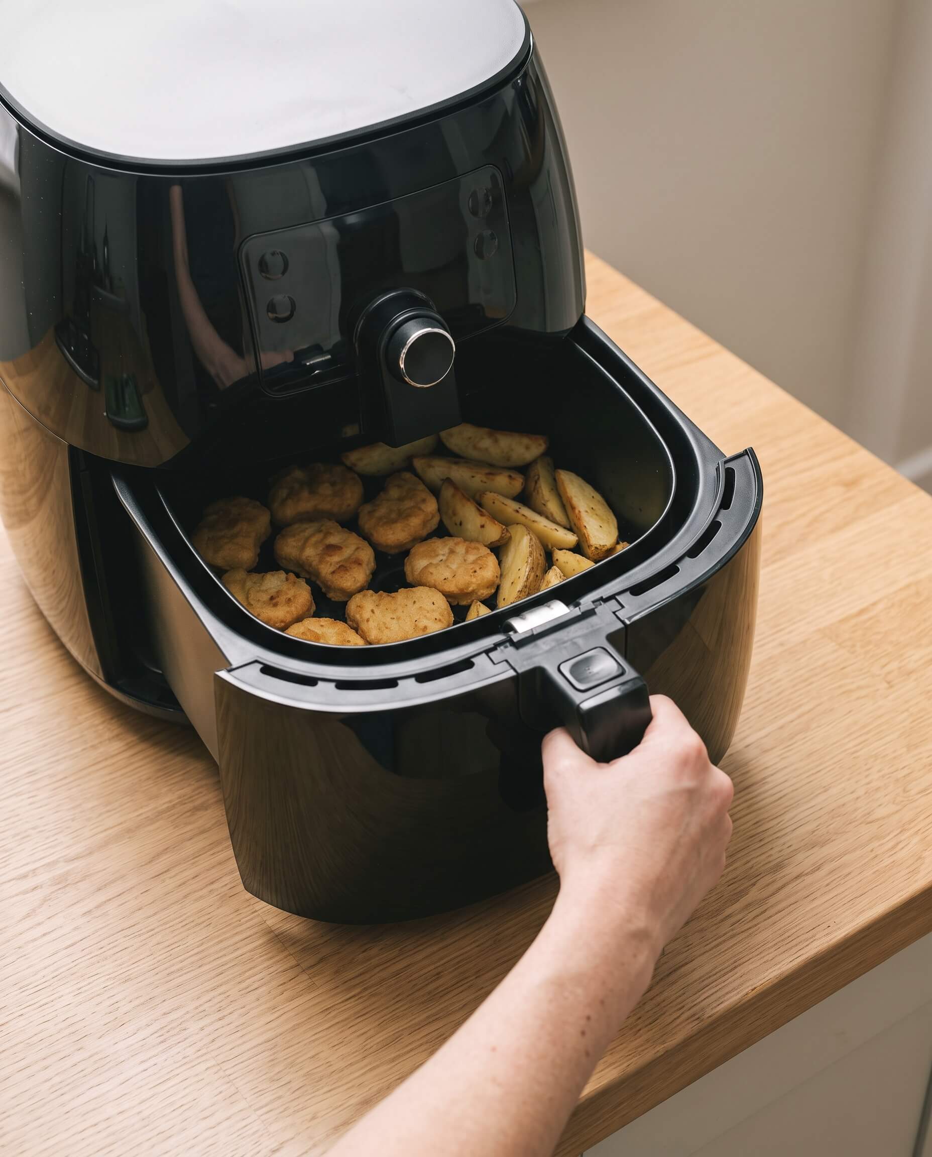 Main qui sort le tiroir d'un air fryer à mi-cuisson, nuggets et pommes de terre à l'intérieur