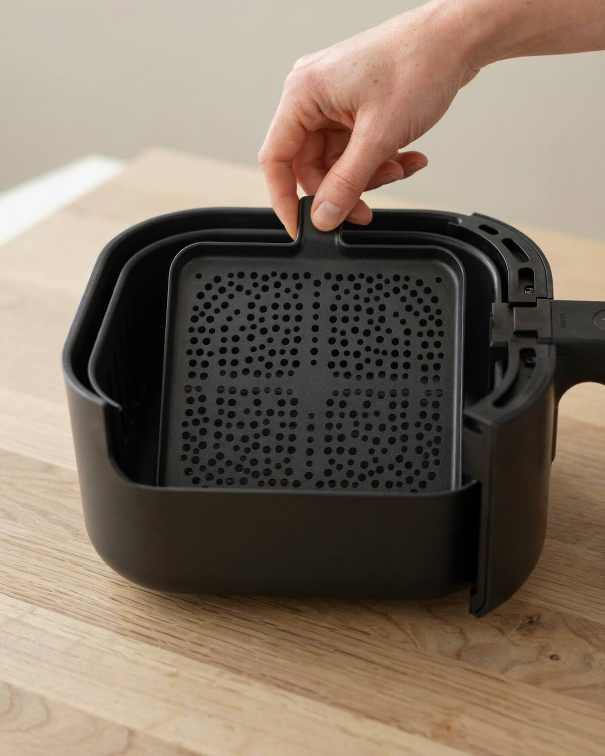 Main qui soulève par sa petite poignée la grille perforée d'un panier d'air fryer vide