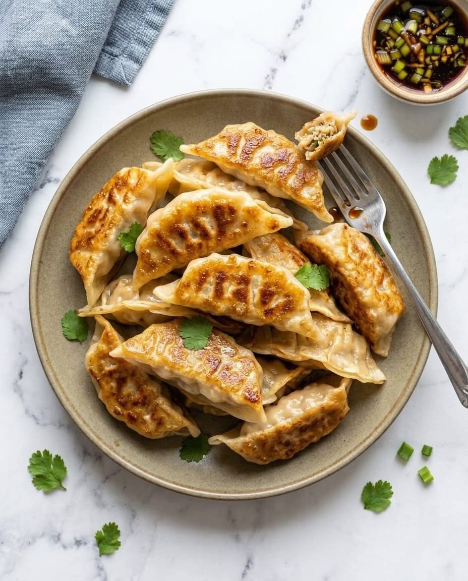 Gyozas dorés sur une assiette, l’un entamé sur la fourchette, feuilles de coriandre et sauce soja aux oignons verts à côté