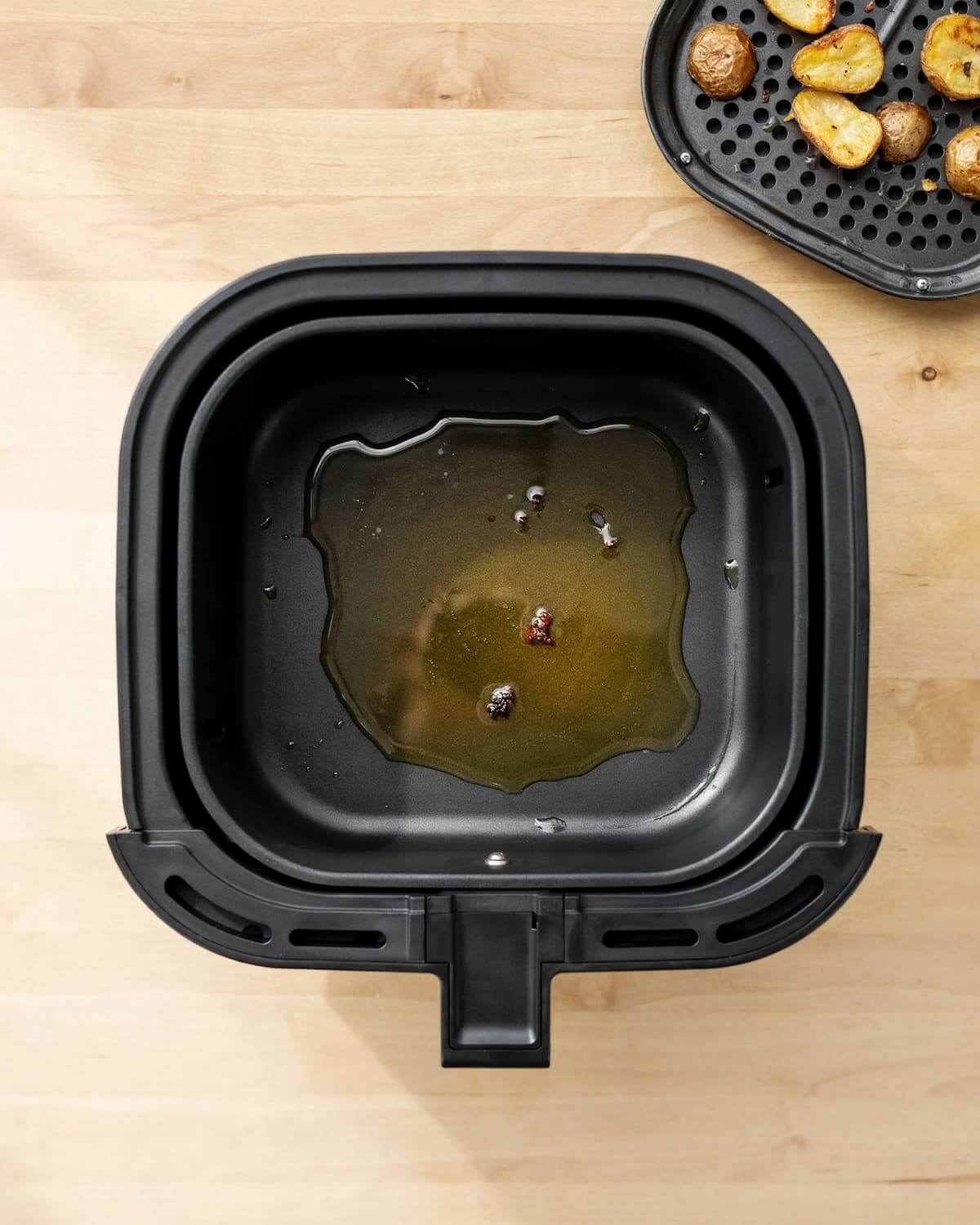 Tiroir d’air fryer, grille retirée, une flaque d’huile en trop au fond avec des miettes brûlées