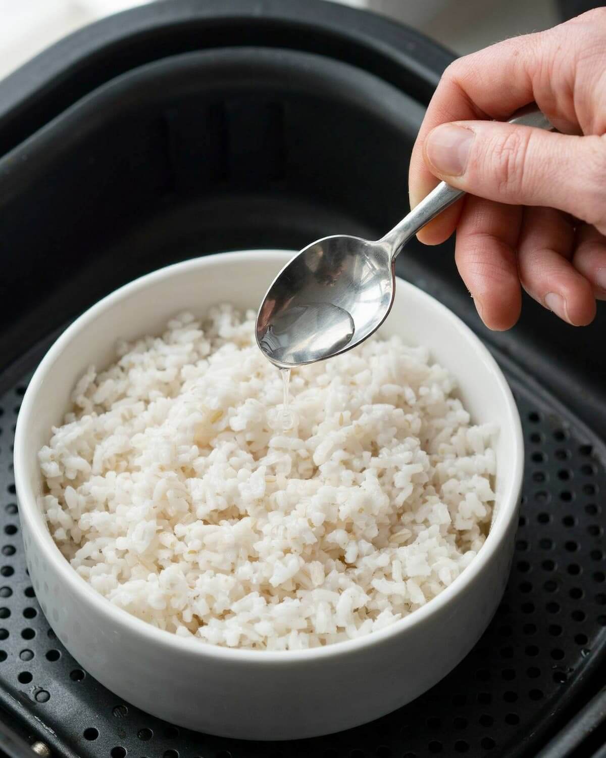 Bol de riz cuit dans le panier d'un air fryer, arrosé d'un filet d'eau à la cuillère avant d'être réchauffé