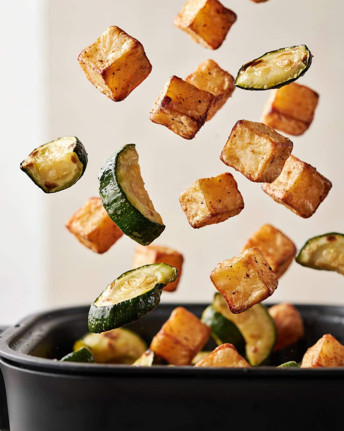 Dés de pommes de terre dorés et demi-lunes de courgette qui sautent au-dessus du panier d'un air fryer qu'on secoue